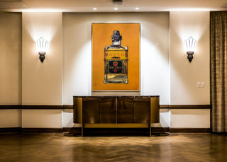 Gemälde von Dominik Lommer zeigt eine Bacardi Rumflasche vor orangefarbenem Hintergrund, hängt über einem Sideboard in einem eleganten Raum mit Wandlampen