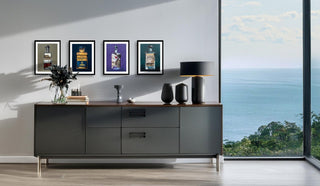 Vier gerahmte Kunstdrucke von Alkoholika-Artefakten von Dominik Lommer hängen über einem modernen Sideboard in einem Raum mit Meerblick.