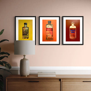 Drei gerahmte Kunstdrucke in rot, gelb, orange aus der Serie „Alcoholica Artefacts“ mit den Motiven Hennessy, Veuve Clicquot und Absolut Vodka hängen nebeneinander an einer Wand über einer Holzkommode mit Tischlampe. Die farbenfrohen Wandbilder eignen sich perfekt als stilvolle Dekoration für die Wohnung oder als originelle Geschenke für Männer.
