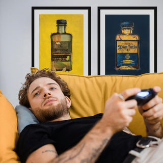 Mann liegt auf gelbem Sofa und spielt Videospiele, im Hintergrund zwei gerahmte Kunstdrucke von Dominik Lommer mit Hennessy- und Don Julio-Flasche