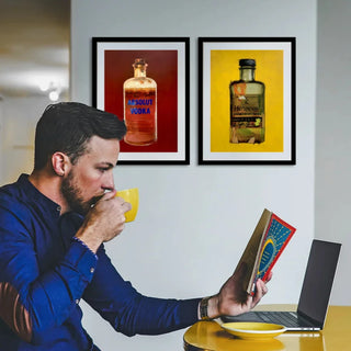 Mann trinkt aus gelber Tasse und liest Buch vor Laptop, im Hintergrund zwei Kunstdrucke von Dominik Lommer mit Alkoholika-Motiven Absolut Vodka und Hennessy, Wandbilder für Wohnzimmer Poster